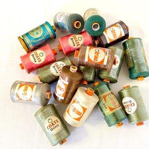 Pode incluir: Uma coleção de carretéis de linha de costura Coats Drima sortidos em várias cores, incluindo tons de verde, marrom, rosa e bege. Cada carretel cilíndrico apresenta uma etiqueta de papel com o nome da marca e texto impresso em preto e laranja.