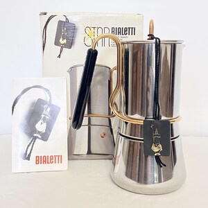 Op de afbeelding: Een roestvrijstalen Bialetti percolator met een goudkleurig handvat en een zwarte hittebestendige greep. De koffiemaker wordt geleverd met de originele witte kartonnen verpakking en een klein instructieboekje, beide voorzien van het Bialetti-logo.