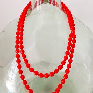 Può includere: Una collana a doppio filo di perline di vetro rosso brillante. Le perline sono rotonde e hanno una finitura sfaccettata. La collana è esposta su una bottiglia di vetro trasparente.