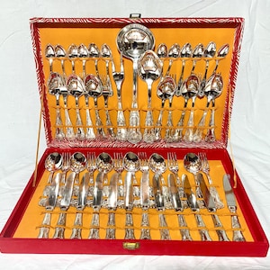 Può includere: Un set completo di posate color argento esposto in una custodia rossa aperta con interno giallo. Il set include cucchiai, forchette, coltelli e utensili da portata. La custodia è rettangolare con coperchio incernierato e manico dorato.