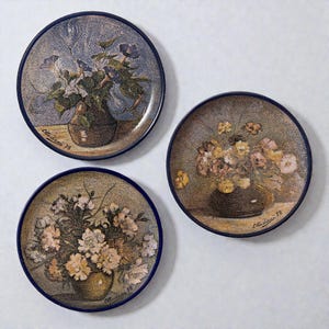 Può includere: Tre piatti decorativi con disegni floreali di natura morta. Ogni piatto presenta un bordo blu scuro e un'immagine centrale di fiori in un vaso. I piatti hanno un'estetica vintage, con la firma dell'artista e l'anno '78 visibili.