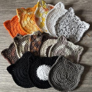 Op de afbeelding: Een set van 10 gehaakte onderzetters in de vorm van een kat in verschillende kleuren, waaronder oranje, geel, wit, bruin, grijs en zwart. De onderzetters zijn rond en hebben de vorm van een kattenoor.