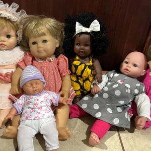 May include: A collection of six dolls featuring various outfits: a peach dress with lace, a pink polka-dot dress, a yellow and black patterned romper, a white and pink floral shirt with white pants, a gray polka-dot dress, and pink overalls.