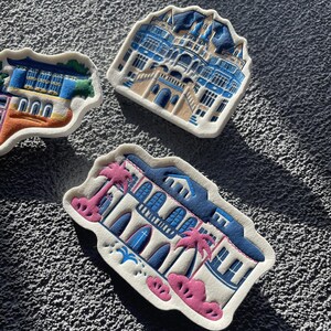 May include: Three colorful ceramic tiles featuring illustrations of buildings. Each tile is shaped like a building and has a white background with blue and yellow details. The tiles are perfect for adding a touch of whimsy to any space.