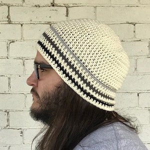 Op de afbeelding: Een witte gehaakte beanie met zwarte en grijze strepen. De beanie wordt gedragen door een persoon met lang bruin haar.