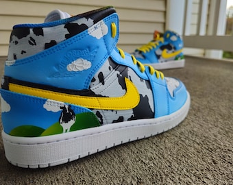 May include: A pair of custom-painted blue and white high-top sneakers with a cow print design, yellow accents, and a green and yellow landscape scene on the side.
