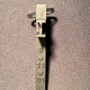 May include: A dark gray metal keychain with the numbers "9", "11", and "01" etched on it. The keychain has a rectangular shape with a small loop at the top for attaching keys. The numbers are in a simple, clear font.