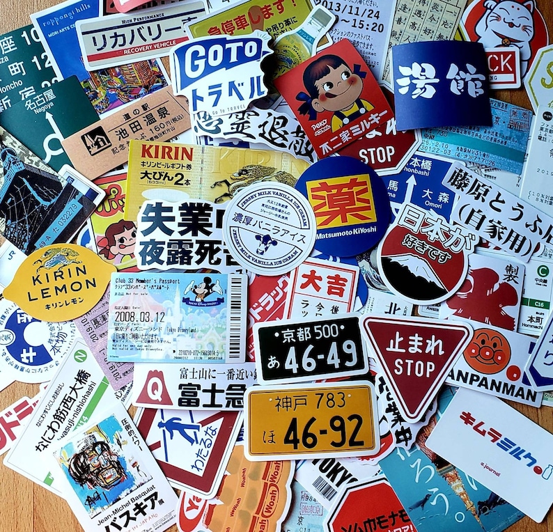 May include: A collection of colorful stickers featuring Japanese characters, illustrations, and phrases. The stickers include images of a cartoon character, a mountain, a stop sign, and a lemon. The stickers are on a brown surface.