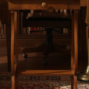 Op de afbeelding: Een bruine houten bijzettafel met een enkele lade met een ronde knop, gebogen poten en een onderste plank. Twee gestapelde boeken met lichtgekleurde ruggen en een wit theekopje op een bijpassend schoteltje staan op het rechthoekige tafelblad.