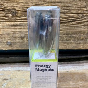Peut inclure: Un emballage en plastique transparent contenant deux aimants énergétiques argentés en forme de goutte d'eau. L'emballage porte une étiquette verte et blanche avec les mots "Energy Magnets". Le fond est une surface en bois.