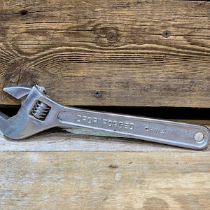May include: A silver adjustable wrench with the words "DROP FORGED CHINA" printed on the handle. The wrench is lying on a wooden surface. The adjustable jaw is open.