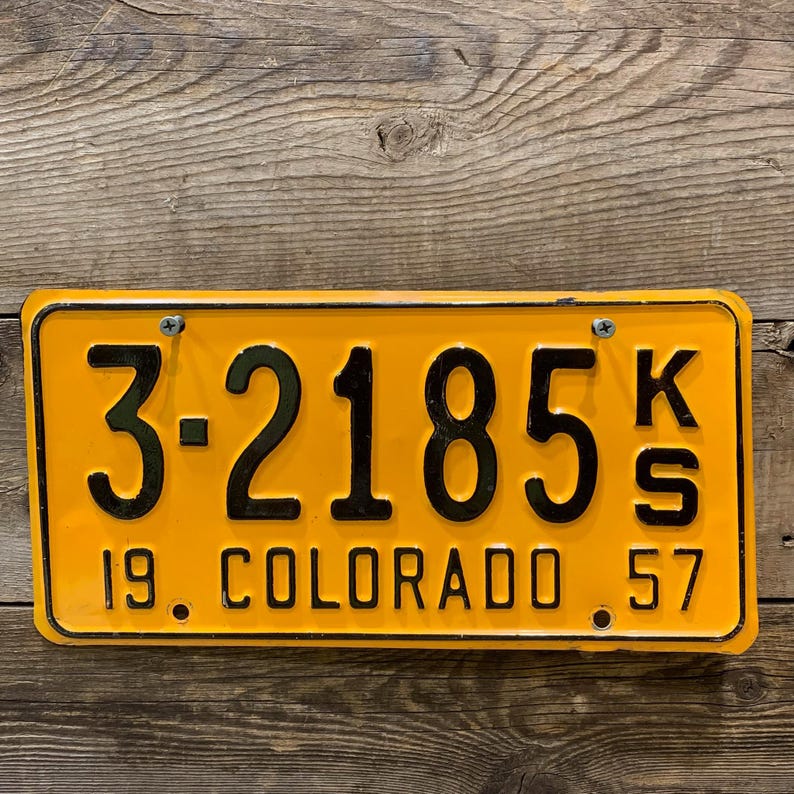 May include: A vintage Colorado license plate with a bright yellow background and black lettering. The plate reads "3-2185 KS" and "19. COLORADO 57". The plate is rectangular and mounted on a wooden surface.