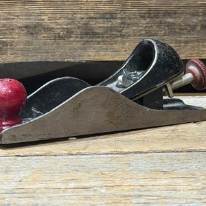 Puede incluir: Un cepillo de mano vintage con base de madera y cuerpo de metal negro. Cuenta con un pomo rojo y una rueda de ajuste roja. La herramienta está diseñada para carpintería y se encuentra sobre una superficie de madera.