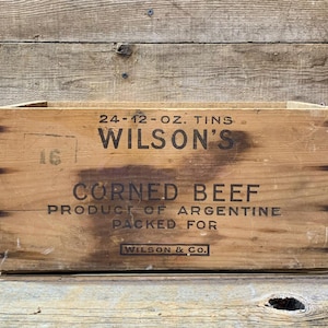 Könnte beinhalten: Eine verwitterte Holzkiste mit dem Text "WILSON'S CORNED BEEF" in schwarzer Schrift. Die Kiste aus hellbraunem Holz zeigt auch "PRODUCT OF ARGENTINE". Metallverschlüsse halten die Kiste zusammen.
