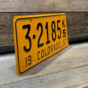 May include: Vintage Colorado license plate with black lettering and numbers on a yellow background. The plate reads "3-2185 KS 19 COLORADO 57". The plate is slightly angled, showcasing its rectangular shape and aged appearance.