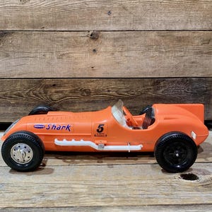 May include: Orange vintage toy race car with black wheels and the text "Shark" and the number "5". The car has a white stripe along the side and a clear windshield. The toy is made of plastic.