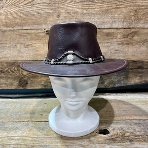 Pode incluir: Um chapéu de couro castanho escuro com aba larga e copa estruturada. O chapéu apresenta uma faixa decorativa com detalhes prateados e um emblema central. O chapéu é exibido em uma cabeça de manequim branca sobre um fundo de madeira.