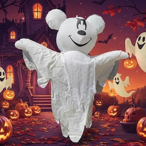 Peut inclure: Une décoration gonflable de fantôme blanc avec une tête ronde, des oreilles noires rappelant une souris et un visage souriant, drapée dans un drap blanc. Le fantôme est placé dans une scène remplie de citrouilles lumineuses et de petits fantômes.