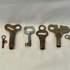 May include: A collection of seven antique keys of varying sizes and designs. The keys are made of metal and show signs of age and wear, with a range of shapes including circular, rectangular, and keyhole-shaped openings. The keys are arranged in a row.