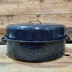 Puede incluir: Una asadera grande de esmalte azul marino moteado con una tapa a juego. La asadera tiene dos asas laterales y un asa en la tapa. El patrón moteado es blanco sobre azul oscuro. La asadera está sobre una superficie de madera.