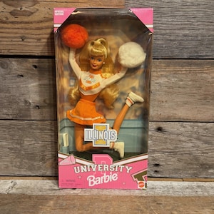 Puede incluir: Una muñeca Barbie edición especial University of Illinois con uniforme de animadora, top naranja y blanco, falda naranja y calcetines blancos. La muñeca sostiene un pompón naranja y uno blanco, con el texto Illinois impreso en la caja.