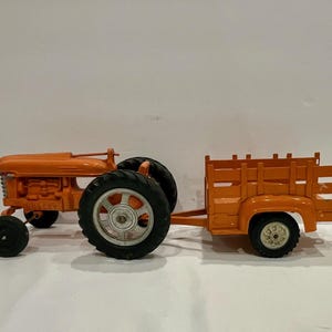 Może przedstawiać: Pomarańczowy traktor zabawkowy z pasującą przyczepą. Traktor ma duże czarne opony i srebrną piastę koła. Przyczepa ma listwową skrzynię. Traktor i przyczepa połączone są metalowym drążkiem.
