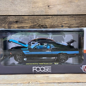 May include: A black 1970 Ford Mustang "Foose" Gambler S14 diecast model car with blue accents, displayed in a clear plastic case. The hood and trunk are open, showcasing the engine and interior details. The model is from M2 Machines.