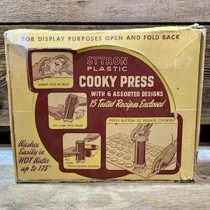 May include: Vintage "Cooky Press" box made of Styron plastic. The box is yellowed with age and features illustrations of the cookie press in use. Text on the box includes "With 6 Assorted Designs" and "15 Tested Recipes Enclosed."