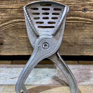May include: A vintage, silver-colored citrus juicer. The juicer has a perforated bowl and two handles that cross. The handles are textured with a dotted pattern. The juicer is made of metal and sits on a wooden surface.