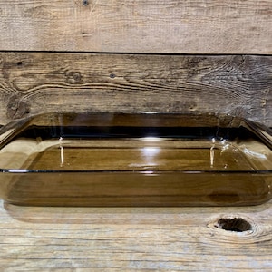 May include: A rectangular, amber-colored glass baking dish with a slightly flared rim and handles. The dish is empty and sits on a wooden surface. The glass has a transparent quality.