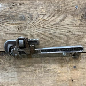 May include: A vintage, well-worn pipe wrench with a dark, metallic finish. The wrench features a toothed jaw, an adjustable screw, and the text "TRADE TRIMONT MARK 10" on the handle. The tool rests on a weathered wooden surface.