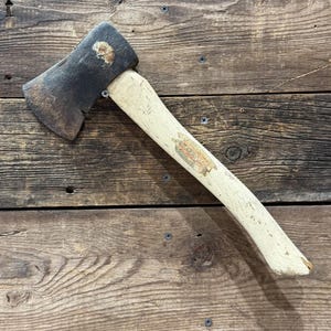 May include: A vintage axe with a weathered, dark metal head and a cream-colored wooden handle. The handle has a faded label. The axe is positioned diagonally on a wooden surface, showcasing its aged appearance and rustic charm.