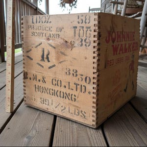 May include: A vintage wooden crate with dovetail joints, featuring stenciled text and graphics. The crate has the words "JOHNNIE WALKER" in red, along with other markings. The crate is made of light-colored wood and appears aged.