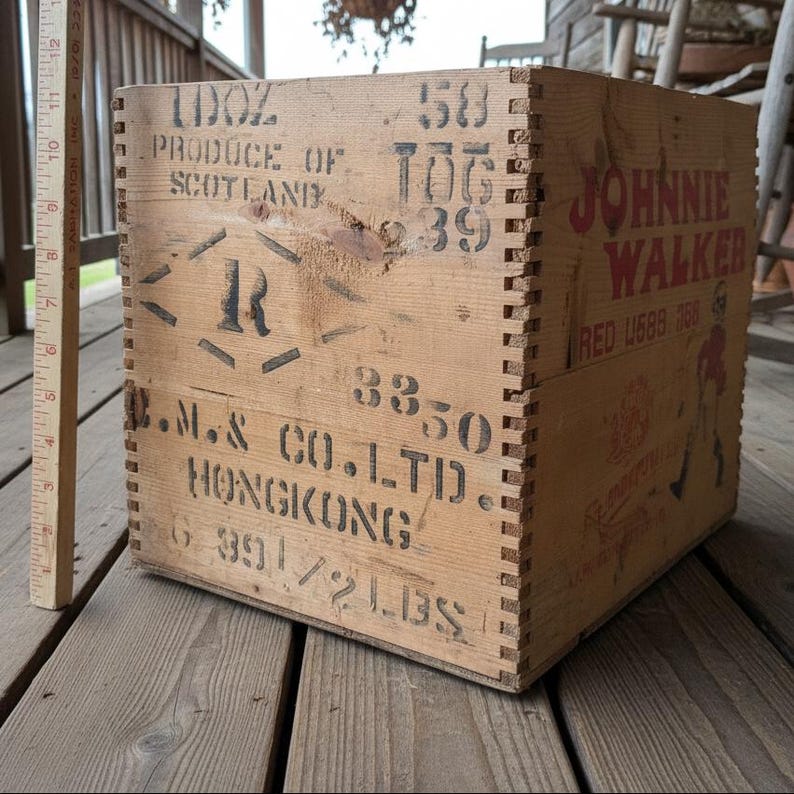 May include: A vintage wooden crate with dovetail joints, featuring stenciled text and graphics. The crate has the words "JOHNNIE WALKER" in red, along with other markings. The crate is made of light-colored wood and appears aged.