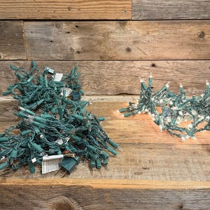 May include: Two strands of string lights with dark green wire and clear incandescent bulbs. One strand is coiled into a dense pile, while the other is partially extended and illuminated, showing the small glass bulbs attached to the wire.