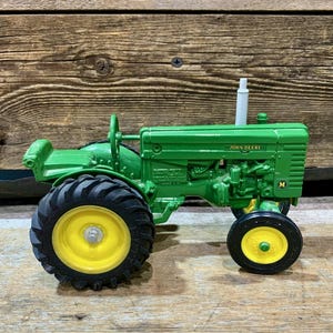Puede incluir: Un tractor de juguete verde con neumáticos negros y llantas amarillas. El tractor tiene las palabras "JOHN DEERE" impresas en el lateral. El tractor es un modelo vintage.