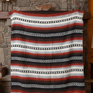 May include: A woven blanket featuring horizontal stripes in shades of rust red, black, white, and light gray. The textile displays repeating geometric patterns in black and white across the surface and includes a fringed edge at the bottom.