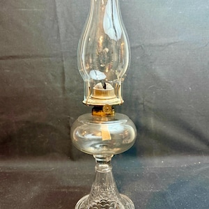 May include: A clear glass oil lamp with a bulbous base and a tall, flared chimney. The lamp has a decorative, patterned base and brass accents. The lamp is designed to hold oil and a wick for illumination.