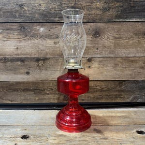 May include: A vintage-style oil lamp featuring a vibrant red glass base with a textured, embossed pattern and a clear, bulbous glass chimney. The lamp includes a metallic burner assembly situated between the red glass reservoir and the chimney.