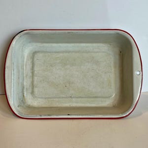 Puede incluir: Bandeja rectangular para hornear con borde rojo e interior blanco. La bandeja tiene esquinas redondeadas y un pequeño agujero en un lado. El interior muestra signos de desgaste.