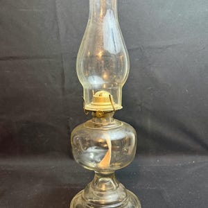 Puede incluir: Una lámpara de aceite de vidrio transparente con base redonda, depósito bulboso y chimenea alta y acampanada. La lámpara tiene un accesorio de metal color latón y una mecha visible en el interior. Diseño vintage, para decoración o colección.