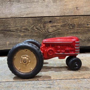 Może przedstawiać: Zabytkowy czerwony traktor zabawka z czarnymi oponami i złotymi kołami. Traktor ma szczegółowy silnik i klasyczny design, prawdopodobnie wykonany z żeliwa. Zabawka jest przedmiotem nostalgicznym.