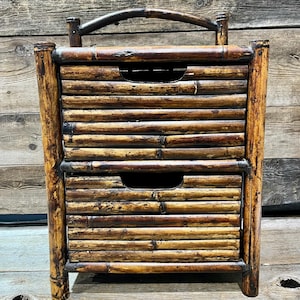 Può includere: Un comodino in bambù a due cassetti con maniglia. Il comodino è costruito con più pali di bambù, creando un aspetto naturale e strutturato. I cassetti hanno ritagli curvi per le maniglie. Il colore generale è un mix di marroni chiari e scuri.