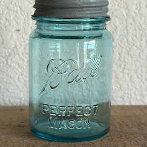 Op de afbeelding: Een vintage blauwe Ball Perfect Mason pot met een grijze metalen deksel. De pot heeft reliëfletters met de tekst "Ball" en "Perfect Mason". De pot is gemaakt van helder blauw glas en staat op een houten oppervlak.