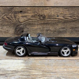 Puede incluir: Modelo de coche deportivo descapotable negro con ruedas plateadas. El coche tiene un diseño elegante con un perfil bajo y un interior visible. El coche está sobre una superficie de madera.