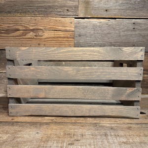 Può includere: Una cassetta rettangolare in legno con una finitura grigia usurata. La struttura è composta da quattro doghe di legno orizzontali sulla parte anteriore, fissate con graffette metalliche visibili alle estremità, creando un design a listelli.