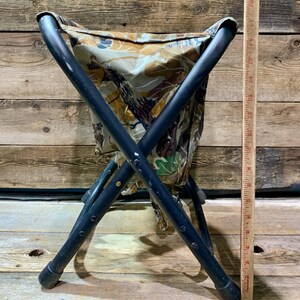 May include: A folding stool with a camouflage seat and black metal frame. The seat fabric features a pattern of brown, green, and tan. The stool is designed with an X-shaped leg structure for stability. A wooden ruler is next to the stool.