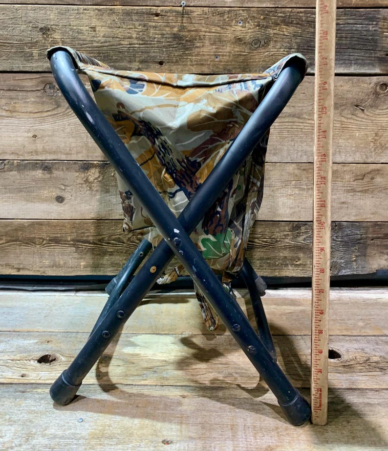 May include: A folding stool with a camouflage seat and black metal frame. The seat fabric features a pattern of brown, green, and tan. The stool is designed with an X-shaped leg structure for stability. A wooden ruler is next to the stool.