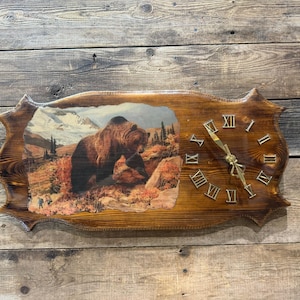 May include: A rectangular wooden wall clock with a scalloped edge and a natural wood grain finish. The left side features a printed image of a brown bear in a mountainous landscape. The right side displays gold Roman numerals and gold clock hands.