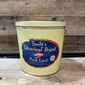 Op de afbeelding: Een vintage gele metalen emmer met een draadhandvat en een deksel. De voorzijde heeft een blauw ovaal etiket met een rode rand, met de tekst Swift's Silverleaf Brand Pure Lard, 16 Pounds Net Weight, en een zegel van het Amerikaanse ministerie van landbouw.